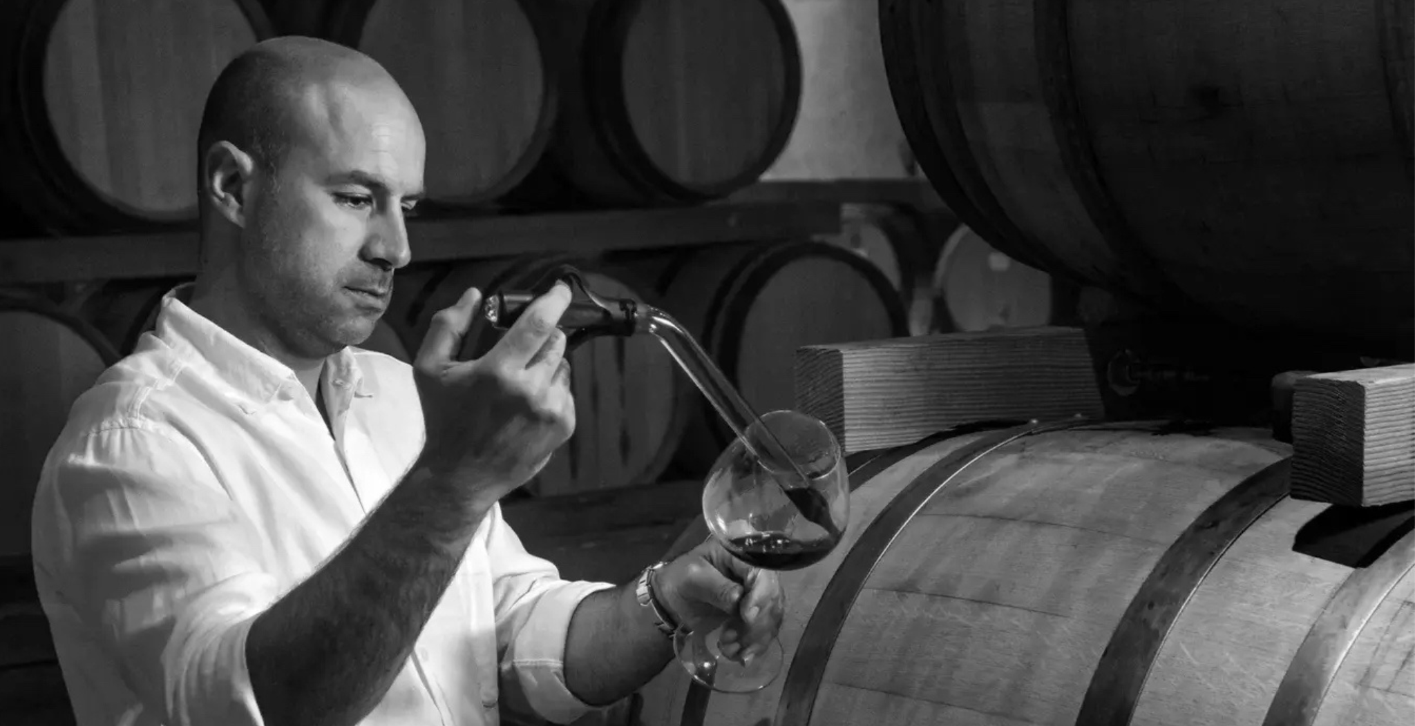 black and white photo of marco surrounded by wine barrels testing a glass of wine